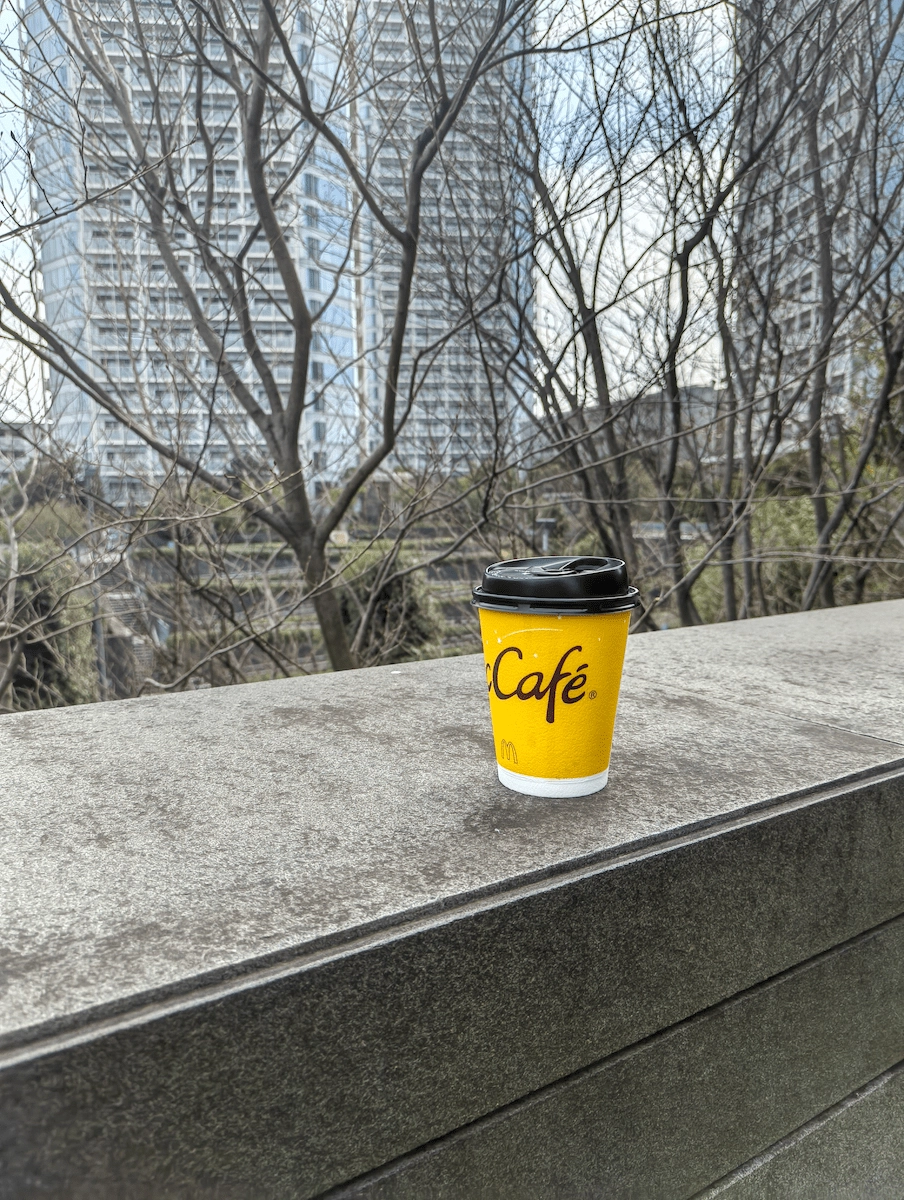 木立の前に置いたマックカフェのカップをPhone(3a) Liteで撮影した背景ボケの雰囲気作例写真