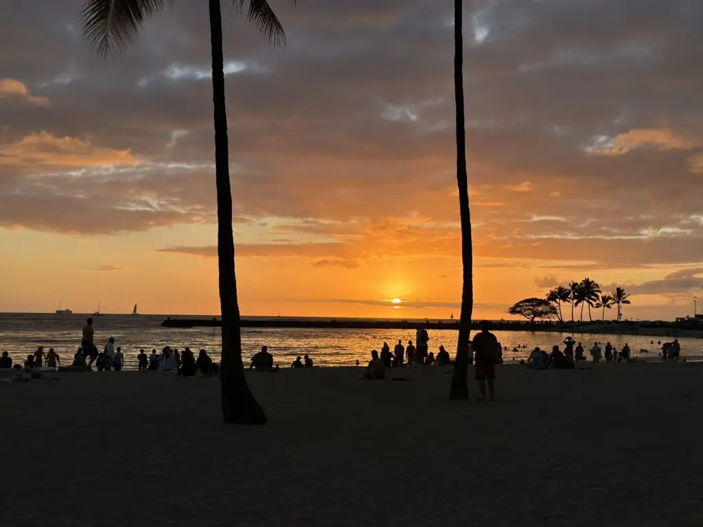 ワイキキビーチの夕景。ヤシの木越しに沈む太陽と、砂浜で夕焼けを眺める人々のシルエット